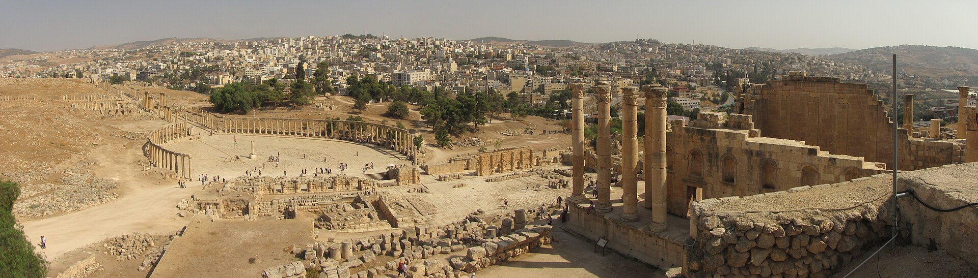 Panorama of Roman ruins at Jerash with a colonnaded oval plaza and a temple, and the modern city behind